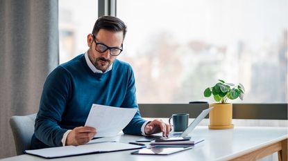 A man looks at paperwork while sitting at his desk with his laptop open nearby.