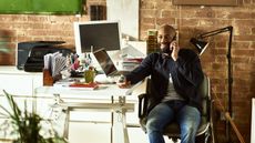 A business owner sits back in his desk chair while he smiles and talks on the phone.