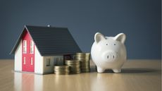 A small model house sits next to three stacks of coins and a white piggy bank.