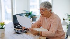 A senior woman uses a calculator in front of a computer.