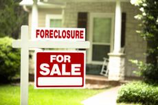 Red and white Foreclosure Home for Sale sign in front of a stone wood house that is for sale and is being foreclosed upon by a financial institution Green grass and bushes indicate the spring or summer season Front porch and windows in background Economic depression recession bankruptcy concepts