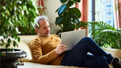 An older man sits on his sofa and looks at his laptop.