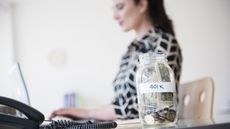 A mason jar labeled 401(k) sits on the desk of a woman working on a computer in the background.