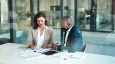 A businesswoman and a businessman look at paperwork at a table in a conference room.